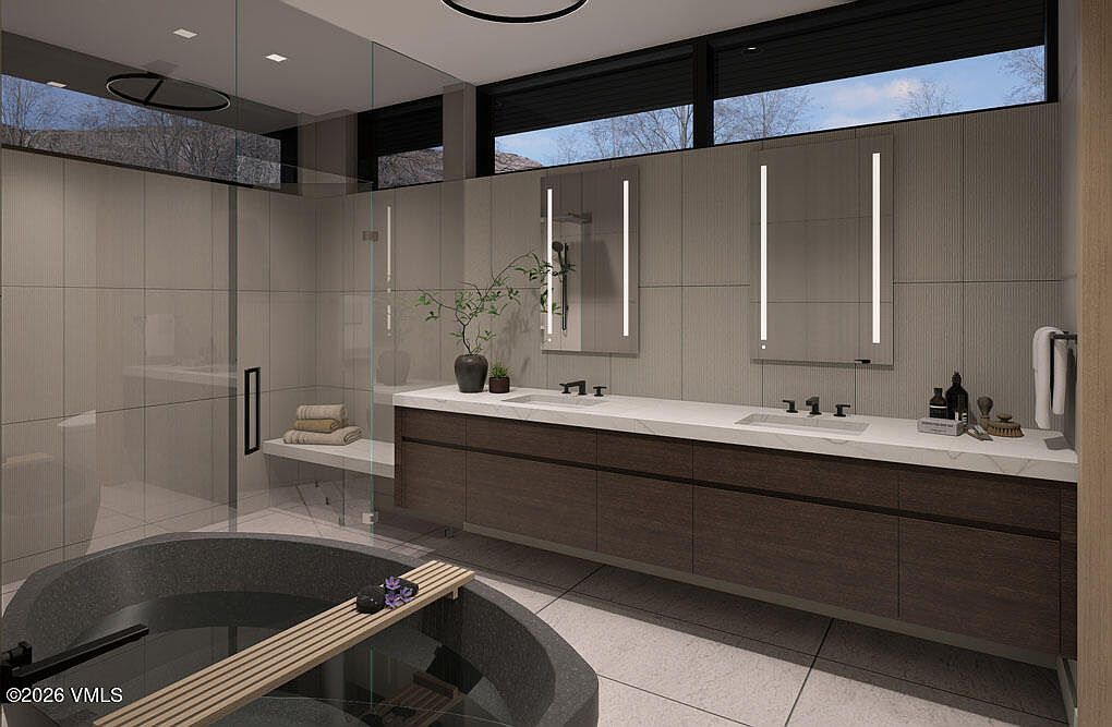 This is a modern primary bathroom featuring a large soaking tub with a wooden plank across it, a double vanity with a white countertop and dark wood cabinets, and a glass-enclosed shower. The bathroom has a minimalist design with clean lines, neutral tile walls, and sleek, illuminated mirrors. The overall impression is luxurious and spa-like.