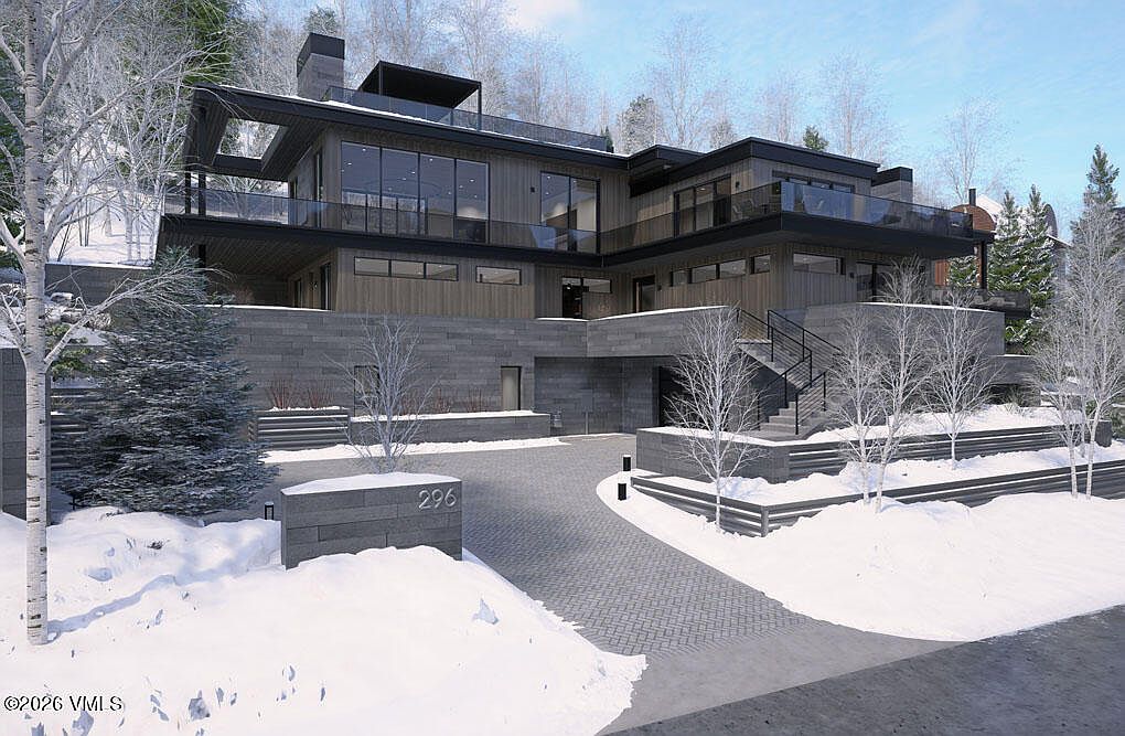 This is a front exterior view of a modern, multi-level home in a snowy, wooded setting. The house features a combination of wood and stone facade, with large windows and balconies offering views of the surrounding landscape. A paved driveway leads to the entrance, and the address '296' is prominently displayed near the driveway entrance.