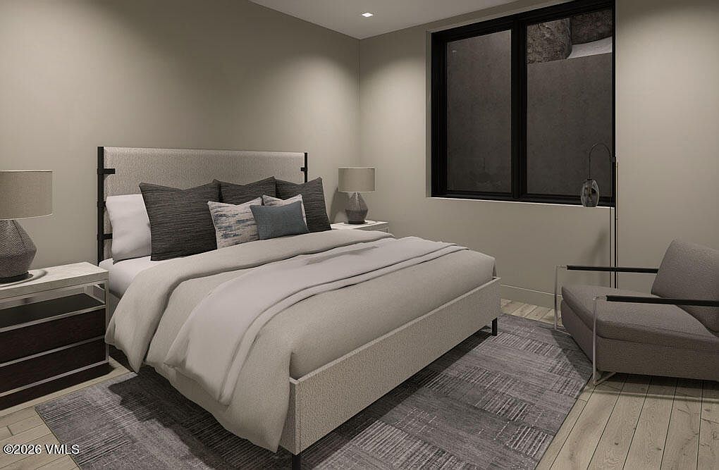 This is a primary bedroom featuring a large bed with a light-colored upholstered headboard and several decorative pillows. The room is decorated in neutral tones, with a gray rug and a modern armchair. A large window provides natural light, and there are bedside tables with lamps on either side of the bed.
