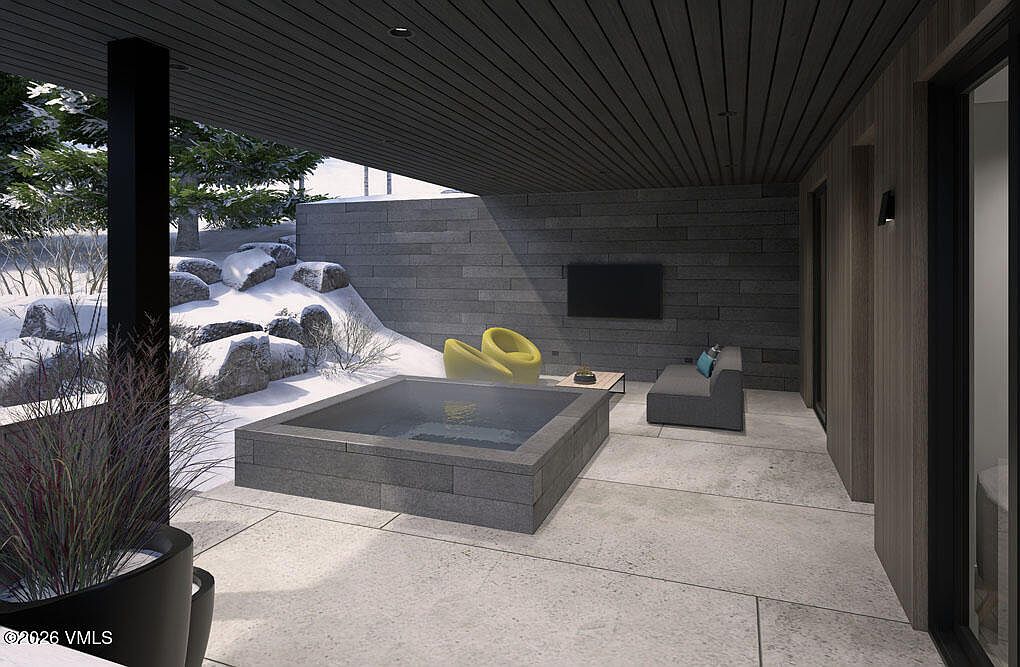 This image showcases a modern patio area with a built-in hot tub, sleek gray stone walls, and contemporary outdoor furniture. The patio features a minimalist design with clean lines and a neutral color palette, creating a sophisticated and relaxing outdoor living space. The snowy landscape visible beyond the patio adds a touch of winter charm.