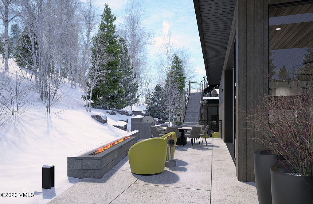 This image showcases a modern patio area with a sleek fire pit, comfortable seating, and an outdoor dining set. The patio is adjacent to a contemporary home with large windows, offering a seamless indoor-outdoor living experience. The snowy landscape in the background adds a touch of serenity and highlights the property's location in a scenic environment.