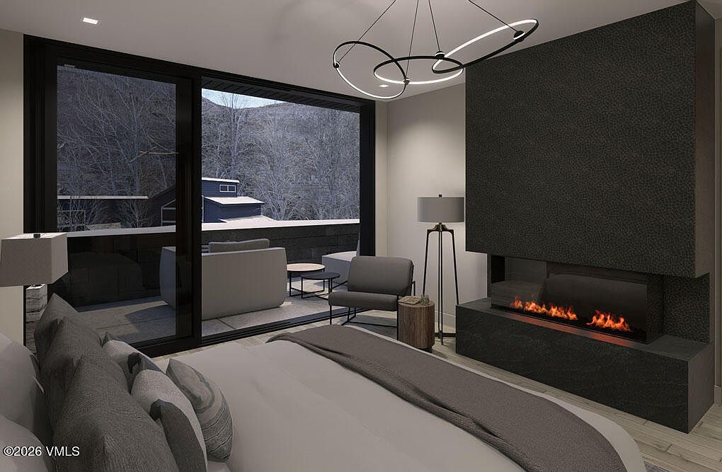 This is a primary bedroom featuring a modern fireplace and a large sliding glass door leading to a balcony. The room is furnished with a bed and a chair, and the decor is minimalist and contemporary. The view from the balcony showcases a snowy landscape, adding to the room's serene and luxurious ambiance.