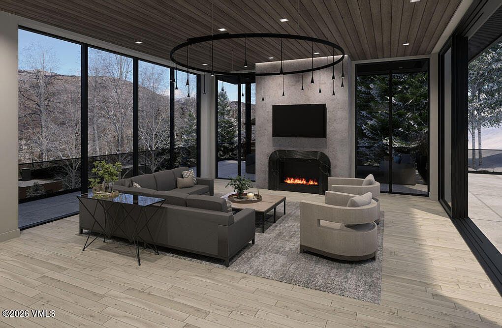 This is a modern living room featuring a gray sectional sofa, two beige armchairs, and a fireplace with a mounted television above it. The room has large windows offering a view of the outdoors, and a unique circular chandelier hangs from the wood-paneled ceiling. The perspective is from a wide angle, showcasing the entire living space and its connection to the exterior environment.