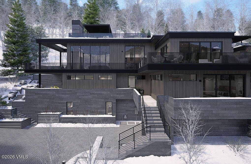 This is a front exterior view of a modern, multi-level home with a sophisticated design. The facade features a combination of wood paneling and stone accents, complemented by large windows and balconies with glass railings. The landscaping includes snow-covered trees and shrubs, creating a serene winter setting.