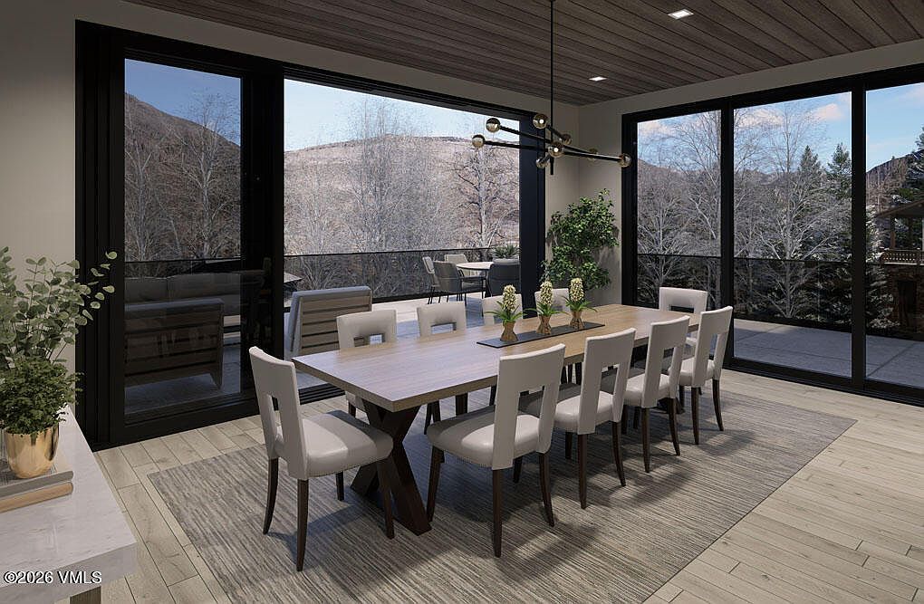This is an interior shot of a dining room featuring a large wooden table with seating for eight. The room has large sliding glass doors that open to an outdoor patio with mountain views. The style is modern and elegant, with a neutral color palette and clean lines.