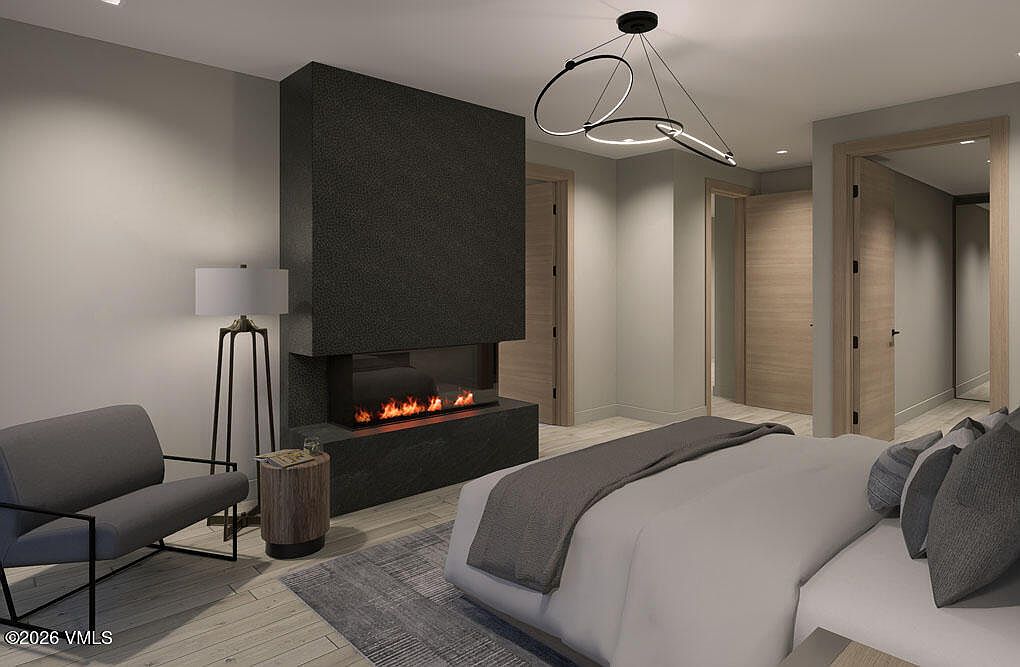 This is a primary bedroom featuring a modern fireplace with a dark stone surround, a gray accent chair, and a contemporary light fixture. The room has a neutral color palette with light wood flooring and doors, creating a serene and sophisticated atmosphere. The bed is neatly made with gray and white linens, and a gray rug adds texture to the space.