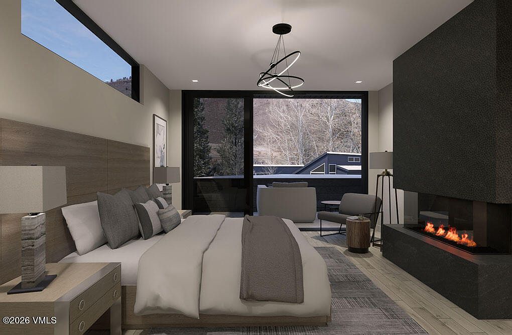 This is a modern primary bedroom featuring a king-size bed with a wooden headboard and neutral-toned bedding. A contemporary fireplace adds warmth, while large sliding glass doors lead to a balcony with outdoor seating. The room is illuminated by a modern chandelier and bedside lamps, creating a serene and luxurious atmosphere.