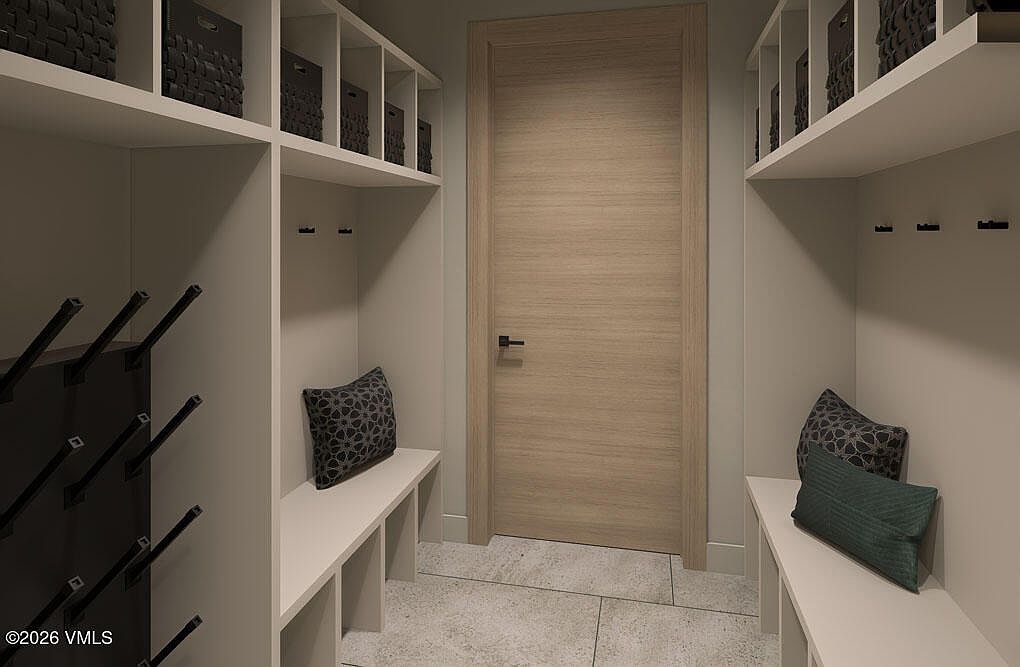 This interior shot showcases a well-organized mudroom or hallway with built-in storage solutions. On either side, there are white shelving units with baskets and hooks, complemented by benches with decorative pillows. A light wood door is centrally located, and the flooring appears to be a neutral tile, creating a clean and functional space.