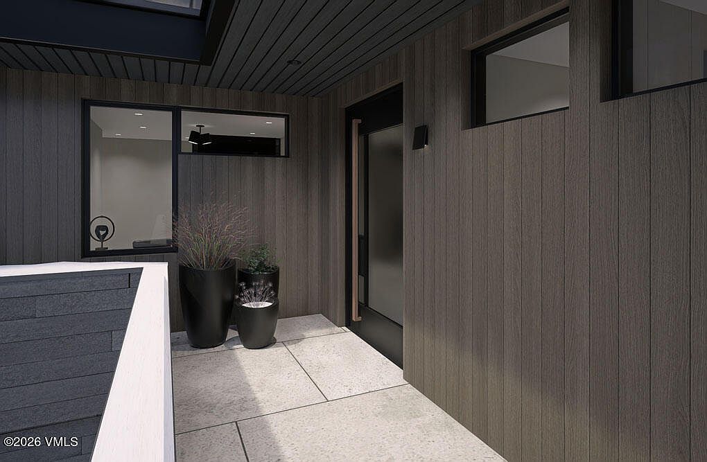 This image showcases a modern entryway with a sleek, dark wood-paneled exterior. The entrance features a black door with a glass panel, complemented by minimalist planters with greenery. The flooring consists of large, light-colored tiles, creating a clean and inviting aesthetic.