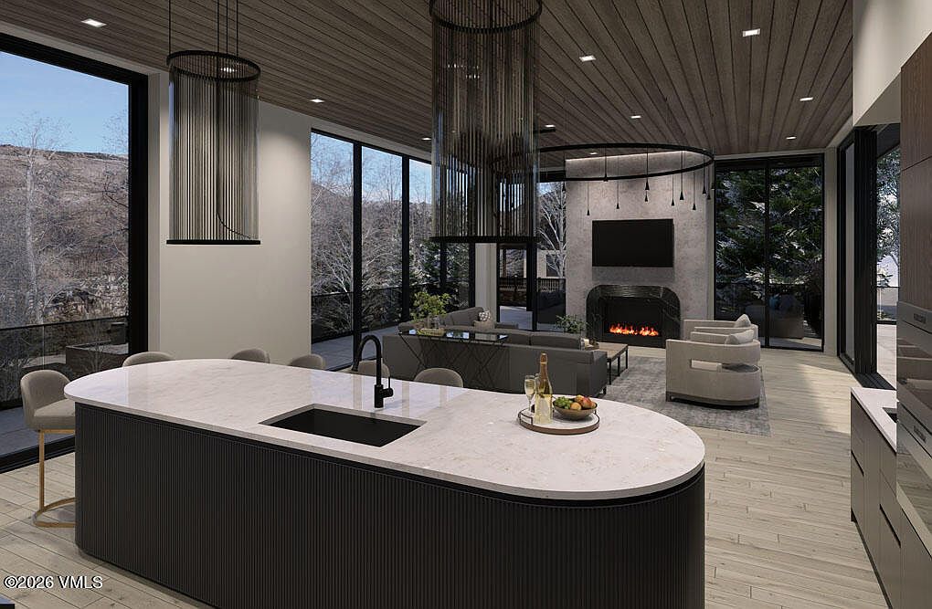 This is a wide-angle shot of a modern, open-concept kitchen and living area. The kitchen features a large, curved island with a white countertop and dark gray base, a black sink, and modern faucet. The living area includes a fireplace, a television, and comfortable seating, all framed by large windows offering natural light and views of the landscape. The overall impression is one of luxury and contemporary design.
