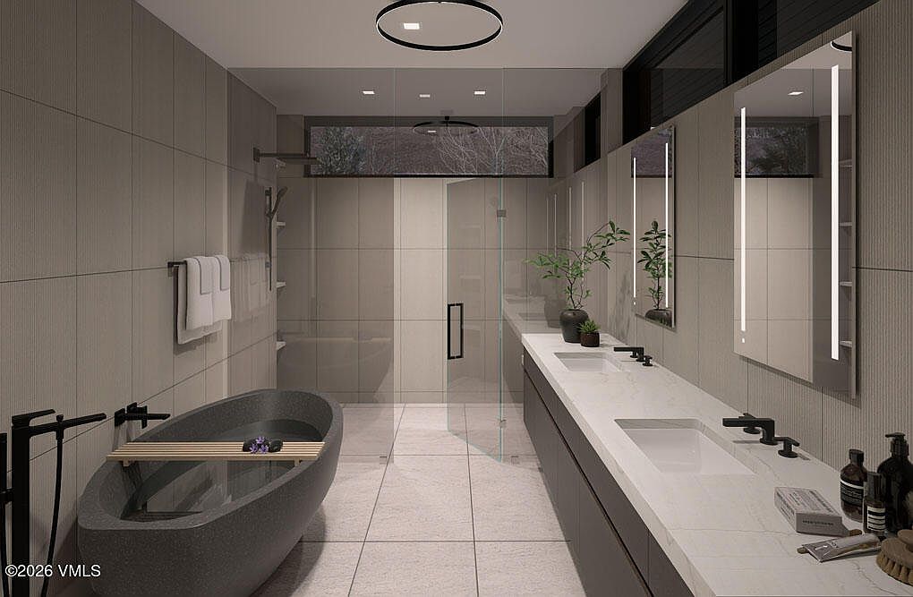 This is a modern primary bathroom featuring a freestanding soaking tub with a wooden shelf, a long double vanity with a white countertop and dark cabinets, and a glass-enclosed shower. The walls are tiled in a neutral tone, and the space is illuminated by recessed lighting and backlit mirrors. The overall impression is clean, minimalist, and luxurious.