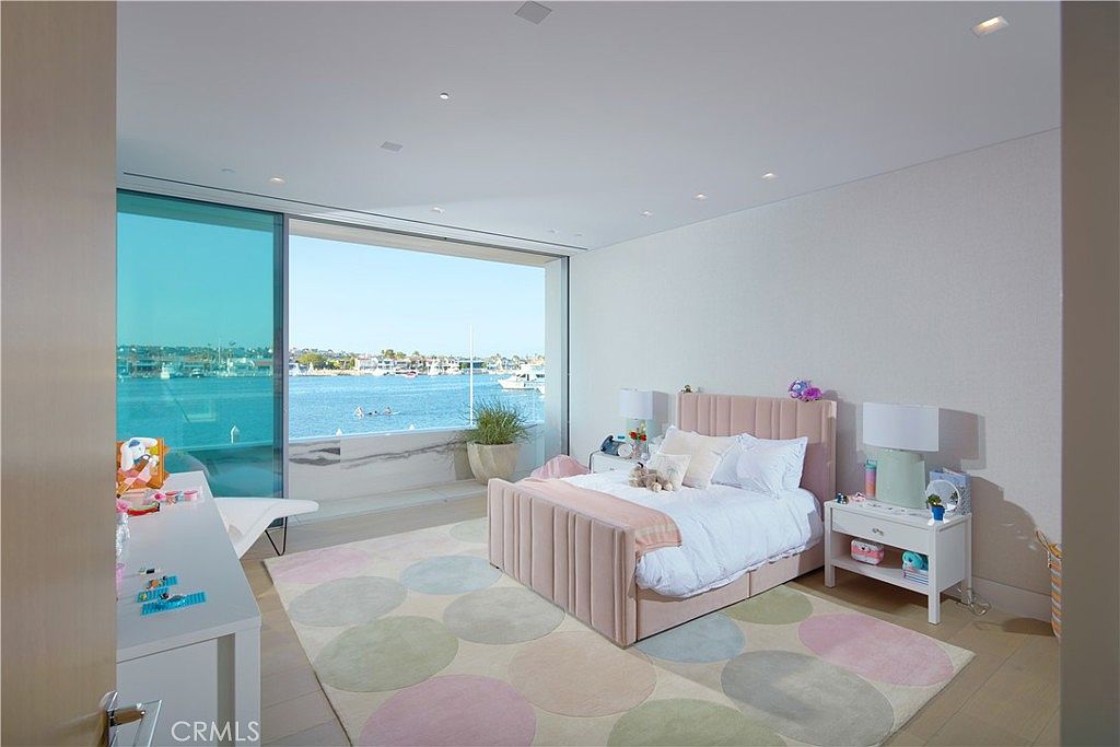This bright and airy bedroom features a soft pink upholstered bed frame and a playful, circular-patterned area rug in pastel tones. A large expanse of floor-to-ceiling glass provides an unobstructed, panoramic view of the harbor and boats outside. The room is furnished with a white desk and a matching nightstand, creating a clean, contemporary aesthetic that emphasizes the waterfront location.