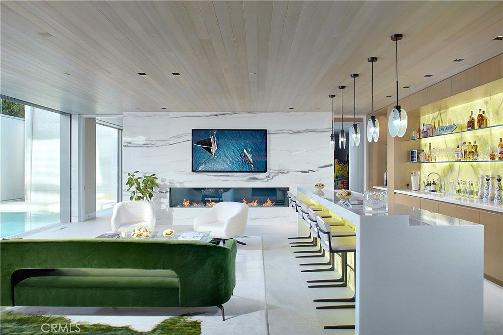 This sophisticated open-concept living space features a striking stone-clad wall with an integrated linear fireplace and a mounted television. A vibrant green velvet sofa anchors the seating area, which is complemented by two white accent chairs and a sleek bar area with illuminated shelving. The room is defined by its light wood-paneled ceiling and expansive glass walls that provide a seamless view of the adjacent pool area.