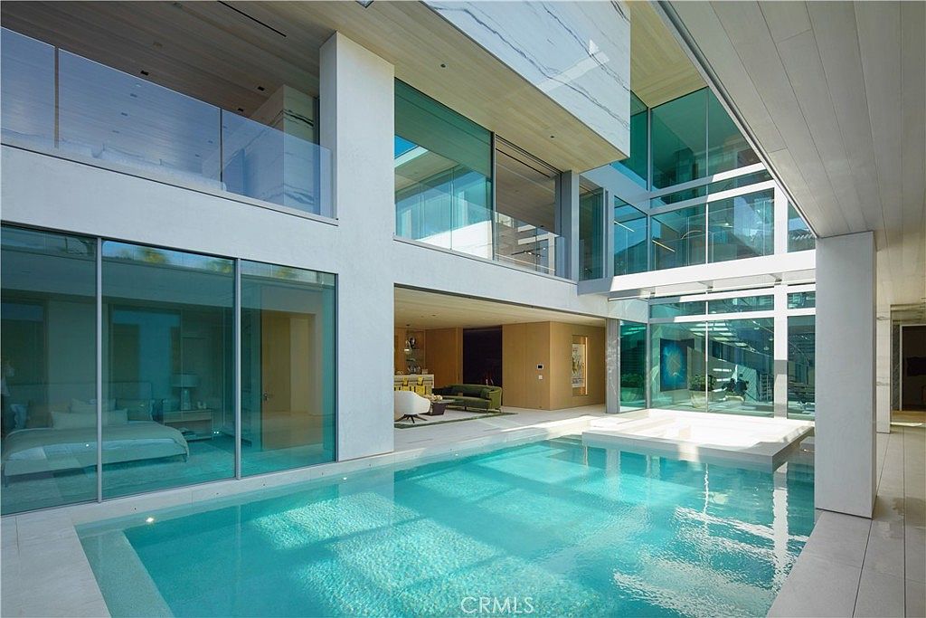 This ultra-modern architectural residence features a central courtyard design centered around a pristine, light-blue swimming pool and integrated spa. The structure utilizes extensive glass walls that create a seamless transition between the indoor living spaces and the outdoor aquatic area, emphasizing a bright, open-concept aesthetic. The clean lines, light-toned surfaces, and minimalist design elements evoke a high-end, resort-like atmosphere.
