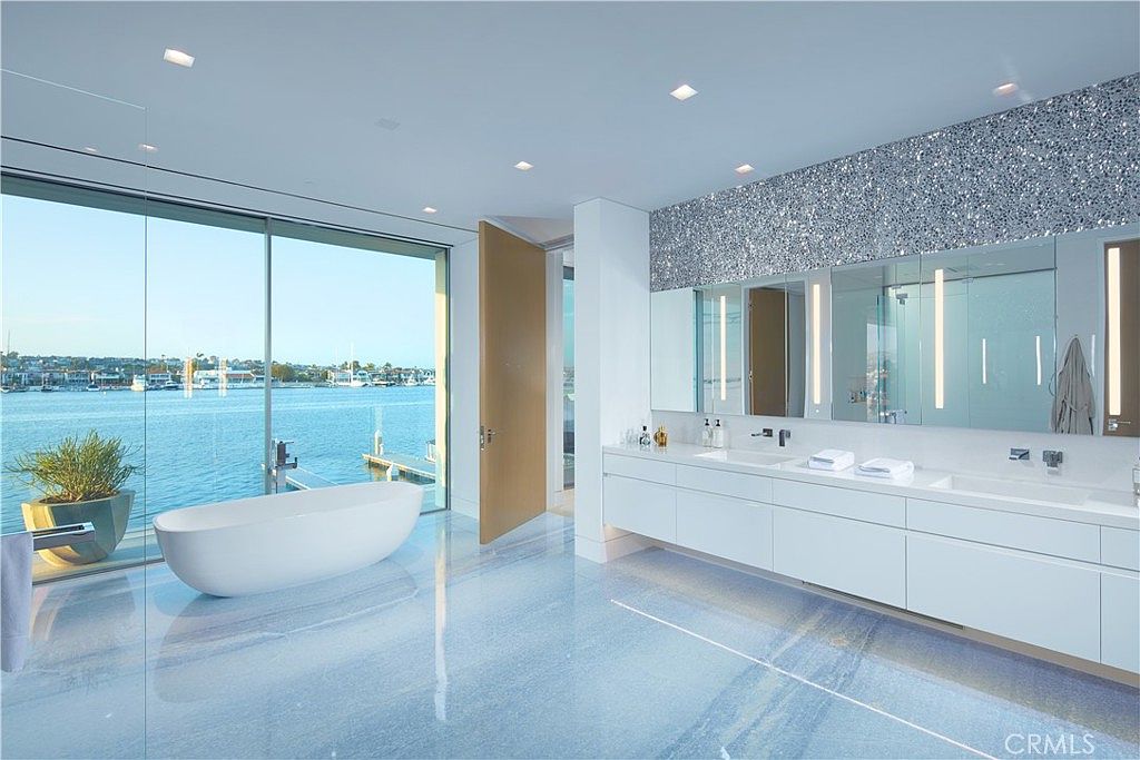 This luxurious primary bathroom features a sleek, freestanding soaking tub positioned next to floor-to-ceiling glass walls that offer expansive waterfront views. The space is anchored by a long, minimalist double vanity with integrated sinks and a striking, textured silver accent wall above the mirrors. The polished blue-toned flooring creates a serene, reflective surface that complements the bright, modern aesthetic of the room.