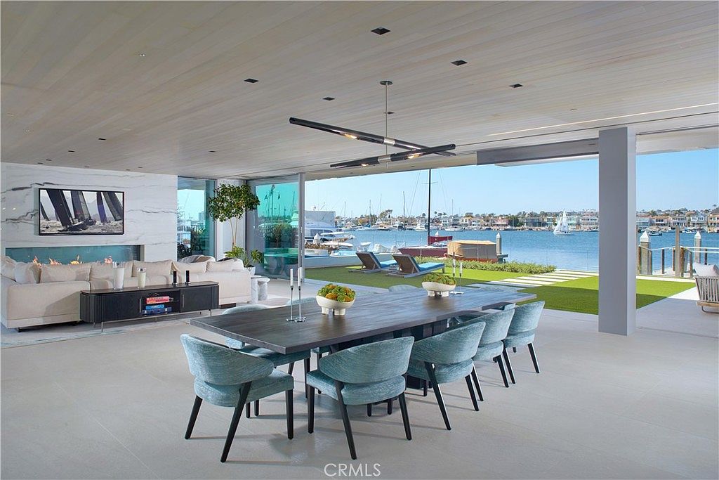 This expansive open-concept living and dining area features a seamless transition to the outdoors through large glass walls that frame a waterfront view. The space is anchored by a long, dark wood dining table surrounded by light blue upholstered chairs, while a plush white sectional sofa sits before a linear fireplace set into a light-toned stone wall. The minimalist aesthetic is complemented by a modern, multi-arm light fixture and a light wood-paneled ceiling, creating a sophisticated and airy atmosphere.