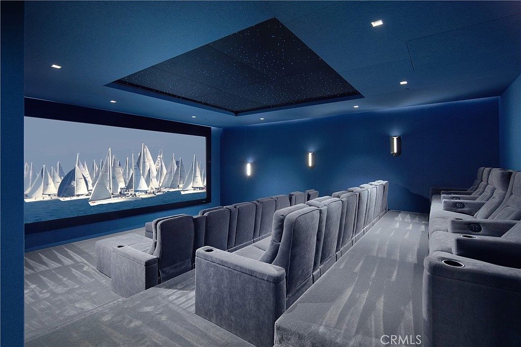 This dedicated home theater features tiered rows of plush, blue velvet seating arranged for optimal viewing of a large projection screen. The room is finished in a deep blue monochromatic palette, accented by a recessed ceiling panel with a starry night light effect and wall-mounted sconces. The tiered layout and professional-grade screen setup create an immersive cinematic experience within the home.