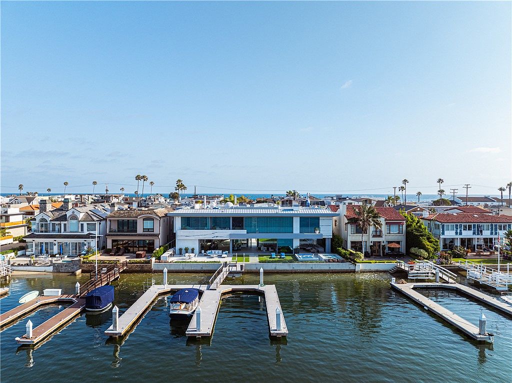 This aerial perspective captures a contemporary waterfront residence situated along a calm bay, featuring a sleek, modern design with expansive glass walls. The property includes a private dock with boat slips, a manicured lawn, and a patio area equipped with a spa. The surrounding neighborhood consists of a mix of traditional and modern homes, all set against a clear blue sky and a distant ocean horizon.