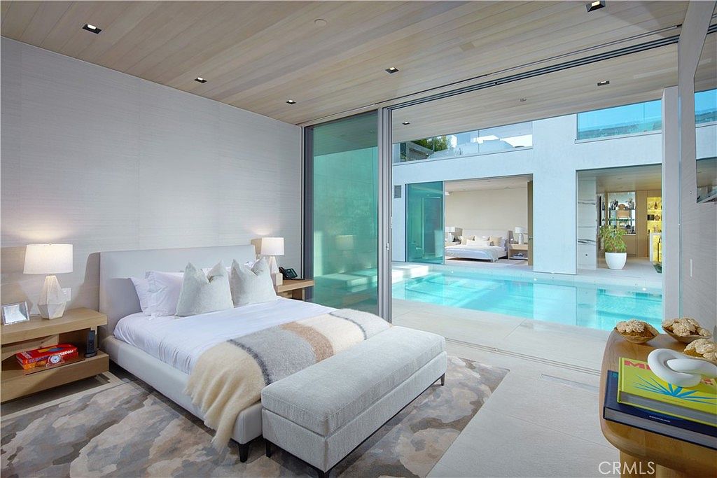 This luxurious primary bedroom features a minimalist design with light wood ceilings and a neutral color palette. Large floor-to-ceiling glass sliding doors open directly onto a private courtyard with a swimming pool, creating a seamless indoor-outdoor living experience. The room is furnished with a plush bed, a matching bench, and a patterned area rug, offering a serene and high-end atmosphere.