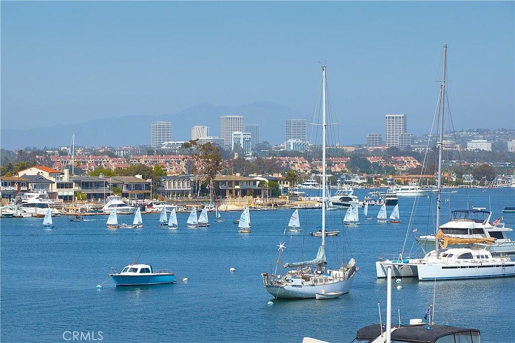 This scenic view captures a bustling harbor filled with numerous sailboats and yachts anchored in calm blue waters. In the background, a coastal residential area features multi-story homes and apartment buildings nestled against a backdrop of distant mountains under a clear sky. The perspective is elevated, likely from a balcony or window, providing a panoramic look at the waterfront lifestyle.
