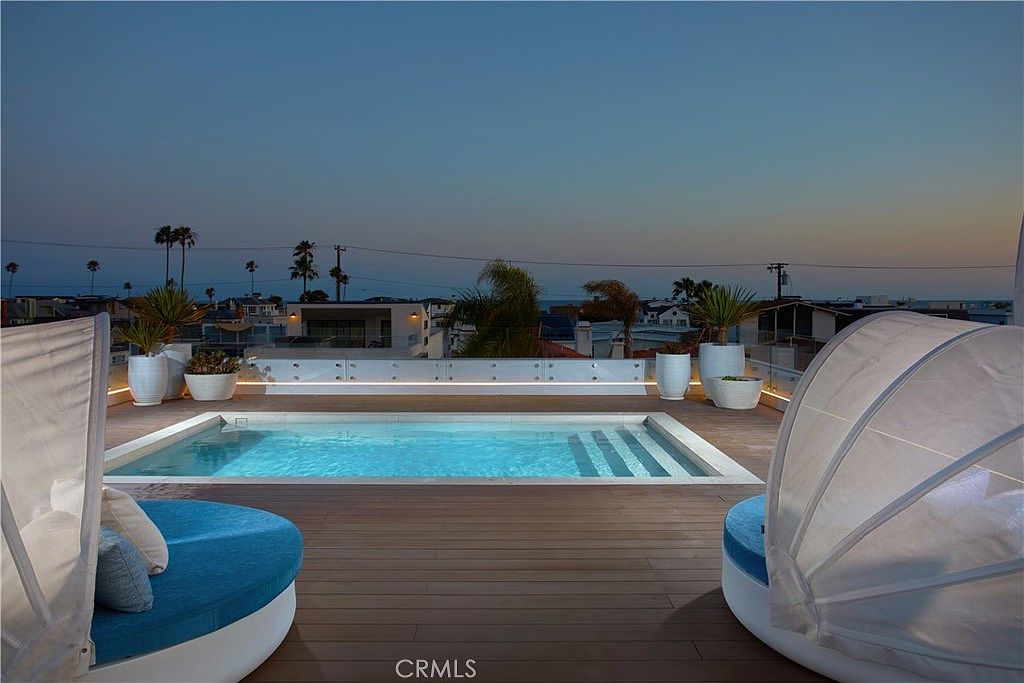 This rooftop deck features a sleek, rectangular swimming pool set into a warm-toned wood-planked terrace. Two circular lounge chairs with white canopies frame the pool, which is bordered by a glass railing that offers an unobstructed view of the surrounding neighborhood and the distant ocean at twilight. The scene is illuminated by subtle, integrated lighting along the base of the glass railing, creating a sophisticated and tranquil atmosphere.