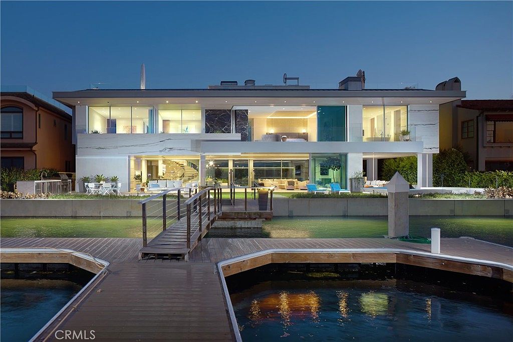 This modern waterfront estate is captured at dusk, showcasing a multi-level architectural design with extensive glass walls that provide a seamless connection between the interior and the outdoor living spaces. The property features a private dock extending into the water, a landscaped patio area with outdoor seating, and a striking spiral staircase visible through the glass. The clean lines, stone-clad exterior, and illuminated interior create a sophisticated and luxurious atmosphere.
