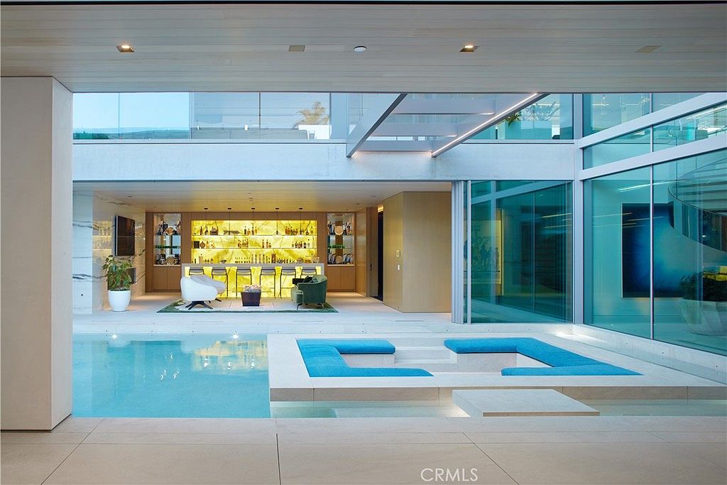 This luxurious indoor-outdoor living space features a sleek swimming pool with an integrated sunken lounge area surrounded by vibrant blue cushions. Beyond the pool, a sophisticated bar area with illuminated shelving and modern seating creates an inviting atmosphere for entertaining. The architecture emphasizes transparency with extensive glass walls that seamlessly connect the interior lounge to the poolside deck.