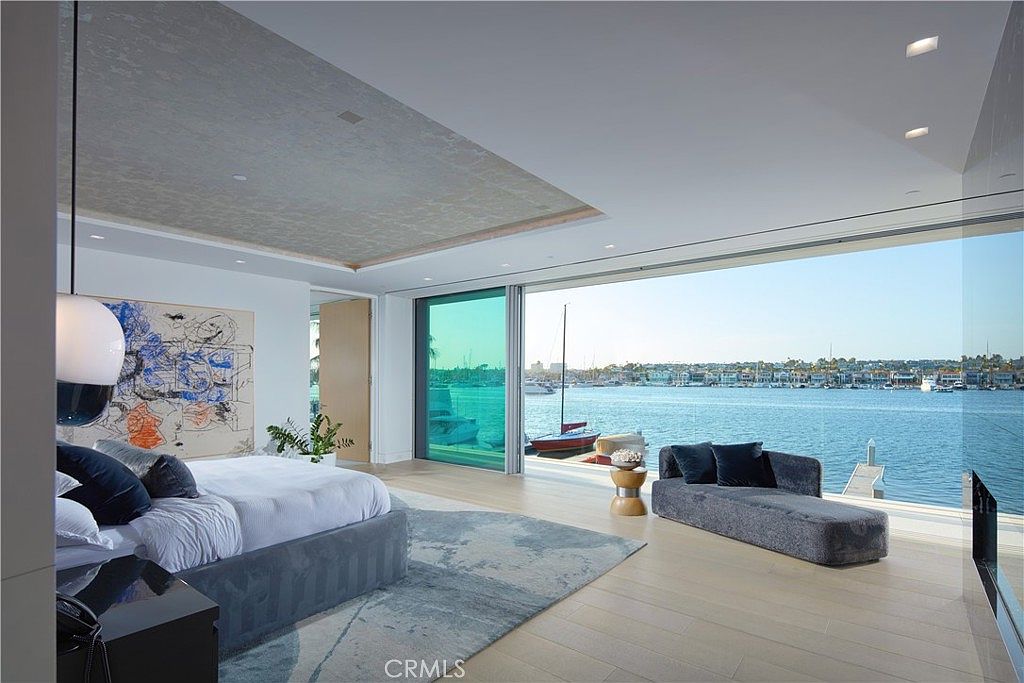 This luxurious primary bedroom features a minimalist design with a large bed and a modern chaise lounge positioned to take advantage of the expansive water views. The room is defined by floor-to-ceiling glass walls that slide open to a waterfront vista, creating a seamless transition between the interior and the harbor. A textured, metallic-toned ceiling detail adds a sophisticated touch to the bright, open space.