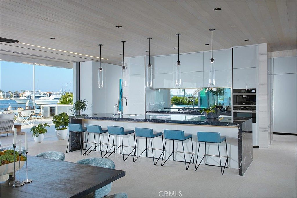 This sleek, modern kitchen features a large island with a dark, veined stone countertop and six vibrant blue bar stools. The space is illuminated by minimalist pendant lights and boasts high-gloss white cabinetry, creating a bright and sophisticated atmosphere. Large glass doors open to a waterfront patio, seamlessly blending the indoor cooking area with the outdoor harbor view.