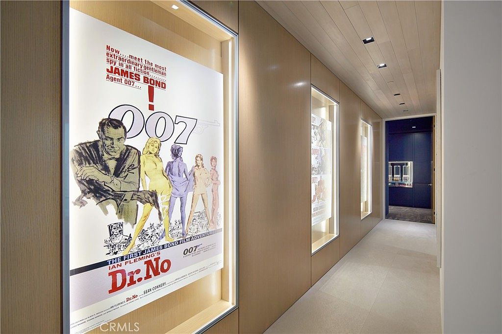 This hallway features a sleek, modern design with light wood paneling on the walls and recessed lighting in the ceiling. The space is accented by illuminated, built-in display cases showcasing vintage James Bond movie posters, creating a sophisticated gallery-like atmosphere. The perspective leads the eye down the corridor toward a dark-toned room at the end, emphasizing the clean lines and contemporary aesthetic of the home.