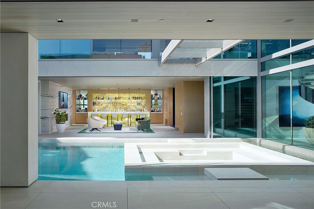 This perspective captures a seamless transition between an indoor bar area and an outdoor pool and spa terrace. The space features a modern, open-concept design with floor-to-ceiling glass walls that blur the lines between the interior lounge and the water features. The pool area is characterized by clean lines, light-toned stone surfaces, and a sophisticated, resort-like atmosphere.