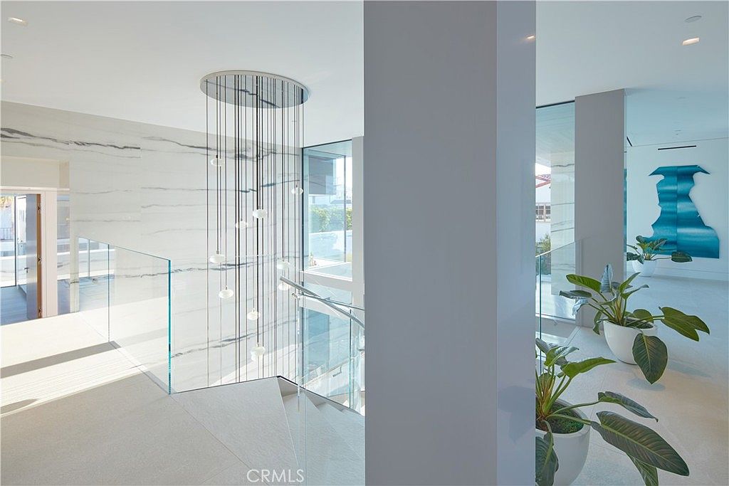 This modern, minimalist staircase area features a striking multi-pendant chandelier suspended over a glass-railed landing. The space is defined by clean lines, white stone wall cladding, and large windows that provide an airy, open feel. Potted plants add a touch of greenery to the bright, contemporary interior, while a vibrant blue abstract artwork hangs on the far wall.
