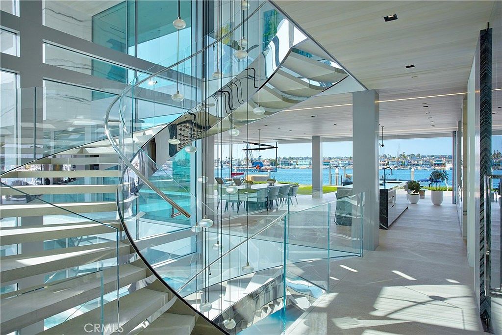 This interior view highlights a striking, modern spiral staircase with glass railings and light-toned treads, serving as a central architectural feature. The open-concept design allows for a clear line of sight through the home to the waterfront beyond, where a dining area and kitchen are visible. The space is bright and airy, characterized by clean lines, neutral tones, and expansive glass elements that emphasize a seamless connection between the indoor living space and the outdoor harbor view.