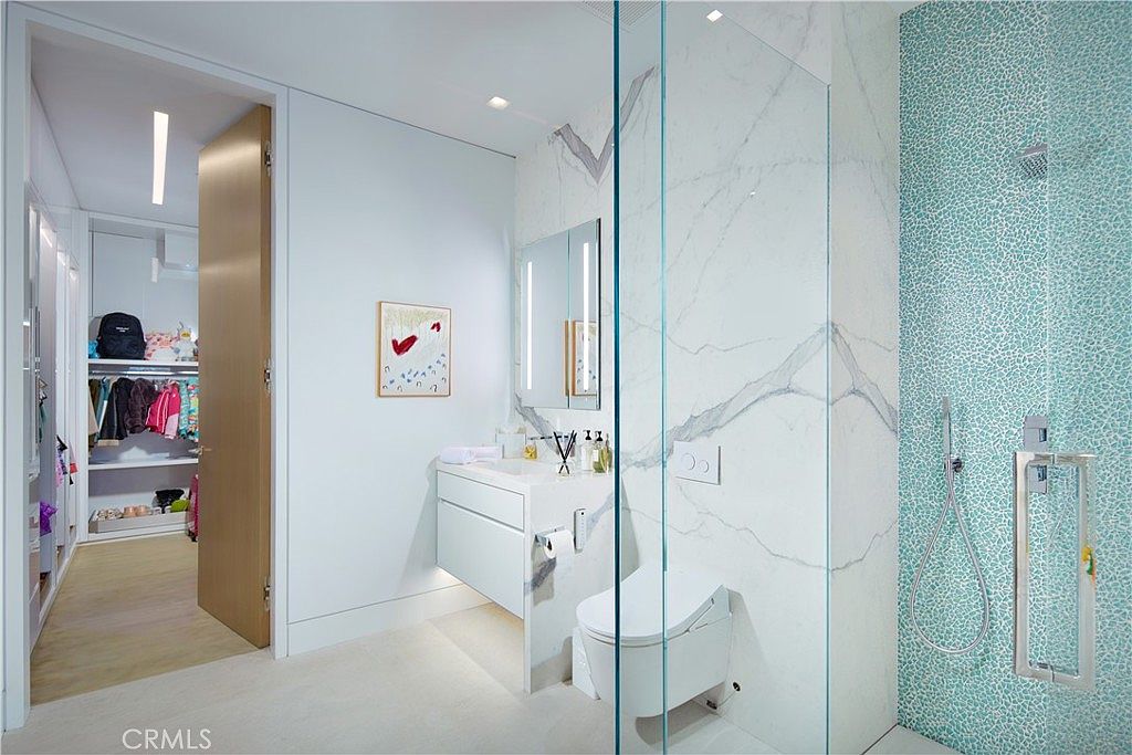 This contemporary primary bathroom features a sleek vanity with a stone countertop and a wall-mounted toilet. A glass-enclosed shower area is accented by a vibrant, textured blue mosaic tile wall, contrasting with the large-format stone wall panels. A doorway leads directly into an adjacent walk-in closet, creating a seamless and functional layout.