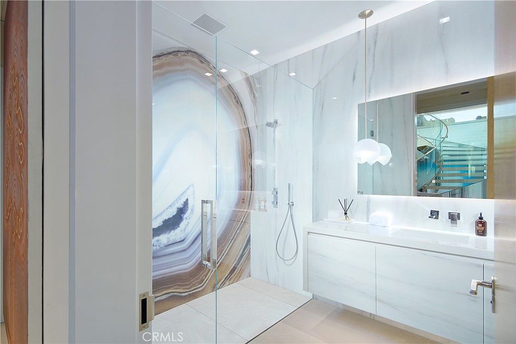 This modern bathroom features a walk-in shower with a striking, large-scale agate-patterned wall panel that serves as a dramatic focal point. A sleek, white floating vanity with a minimalist sink and a large, rectangular mirror sits adjacent to the shower, illuminated by a contemporary pendant light. The space is finished with light-toned stone flooring and walls, creating a clean, high-end aesthetic.