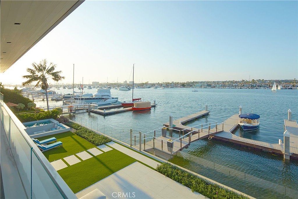 This elevated balcony offers a panoramic view of a serene harbor filled with various boats and yachts. The outdoor space features a manicured artificial lawn, a built-in spa, and lounge chairs, all enclosed by a sleek glass railing. The perspective captures the seamless transition between the private luxury deck and the expansive waterfront setting.
