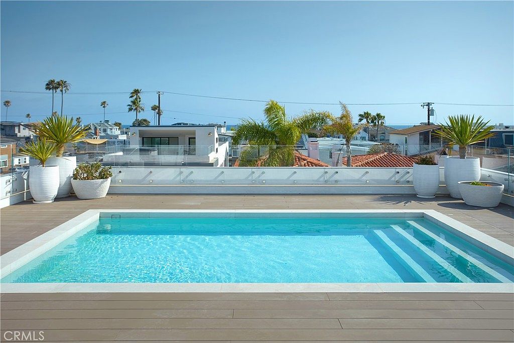 This rooftop deck features a rectangular swimming pool with integrated steps, surrounded by light-toned decking and a glass railing that offers an unobstructed view of the neighborhood and the ocean in the distance. Several large, white planters containing various plants are arranged along the perimeter, adding greenery to the modern, minimalist space. The perspective is eye-level, capturing the serene atmosphere of an elevated outdoor oasis under a clear blue sky.