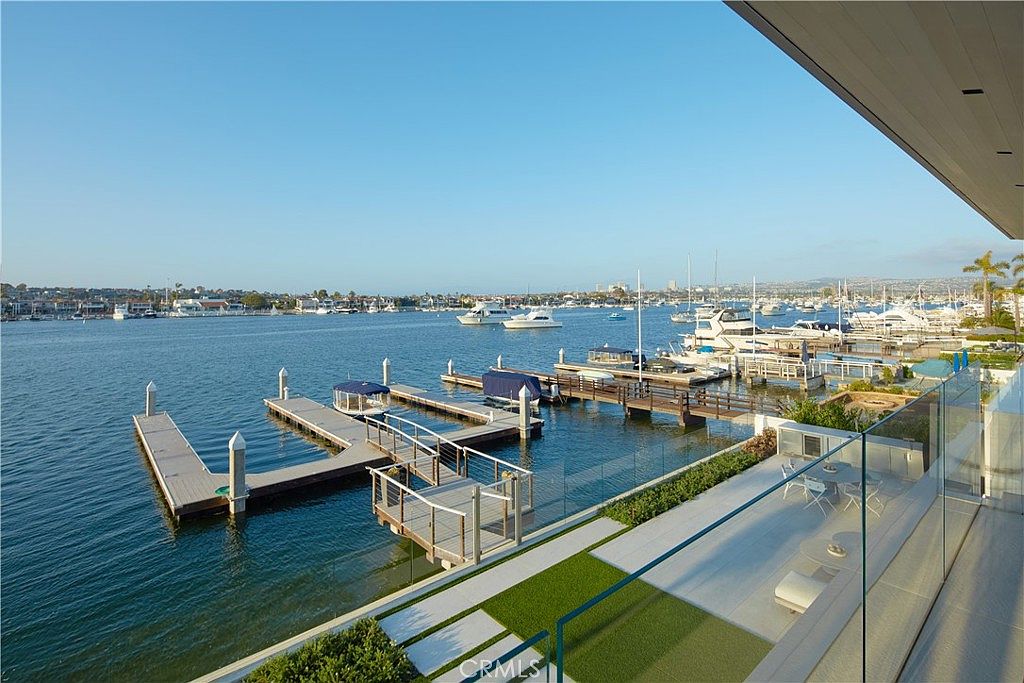 This elevated balcony offers a panoramic view of a serene harbor filled with private docks and various boats. The space features a modern glass railing, a paved patio area with outdoor seating, and a small patch of artificial turf. The perspective is from an upper-level vantage point, capturing the tranquil waterfront setting and the clear blue sky.