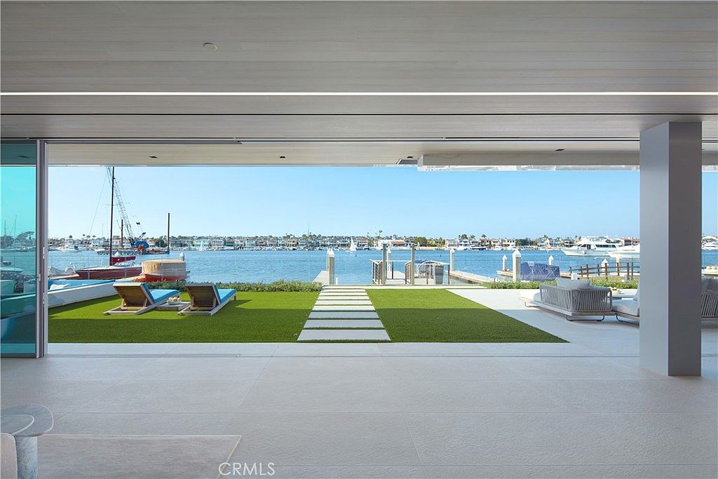This expansive indoor-outdoor living space features a seamless transition to a waterfront patio through massive, retractable glass walls. The interior boasts neutral, light-toned flooring that extends onto the outdoor terrace, which includes a manicured lawn, stone pavers, and lounge chairs overlooking the harbor. The open-concept design emphasizes a bright, airy atmosphere with unobstructed views of the water and docked boats.