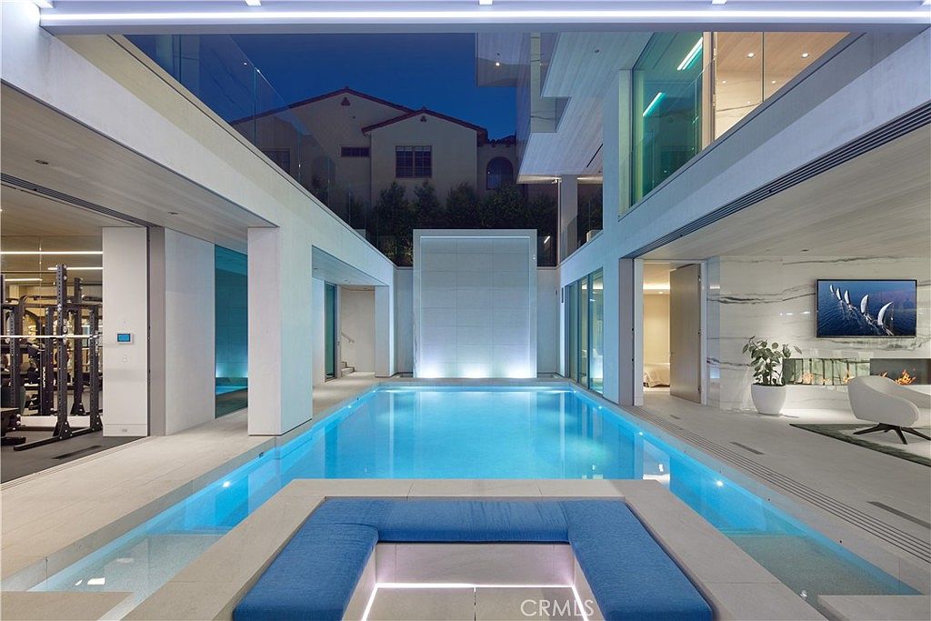 This modern courtyard features a central swimming pool with a sunken lounge area and blue cushioned seating. The space is flanked by a home gym on the left and a living area on the right, both connected by large glass walls that open to the pool. A tall, illuminated water feature serves as a focal point at the far end of the pool, creating a sophisticated and tranquil atmosphere.