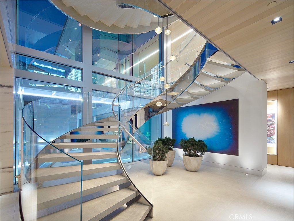 This striking entryway features a dramatic, curved staircase with light-toned treads and a sleek glass railing that winds upward. The space is defined by expansive floor-to-ceiling glass walls that provide a bright, airy atmosphere and views of the exterior. A large, vibrant blue abstract painting serves as a focal point, complemented by potted greenery and a cluster of modern, spherical pendant lights hanging from the ceiling.