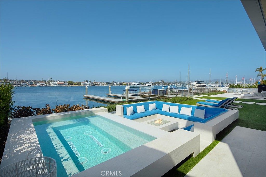 This luxurious waterfront patio features a sleek, rectangular spa adjacent to a sunken lounge area with a built-in fire pit. The space is furnished with bright blue cushioned seating and is set against a backdrop of a calm harbor filled with boats. The modern design emphasizes seamless indoor-outdoor living with a clear glass railing overlooking the water.