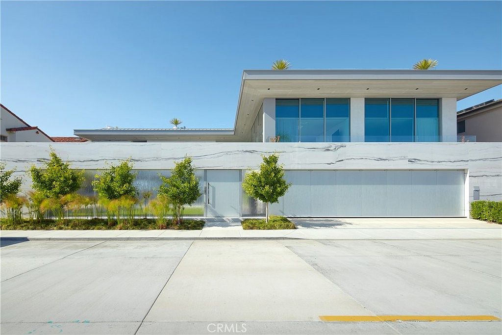 This modern, minimalist residence features a striking facade clad in large, light-colored stone panels with prominent veining. The front elevation showcases a clean, horizontal design with a flat roofline, a central entryway, and a wide garage door integrated into the stone wall. Landscaping includes a row of small, uniform trees and ornamental grasses, set against a clear blue sky, creating a sophisticated and contemporary curb appeal.