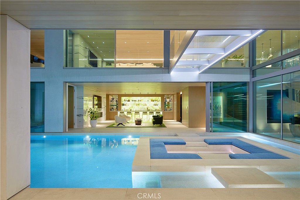 This luxurious indoor-outdoor living space features a large, illuminated swimming pool that extends directly into the home's main living area. A sunken lounge area with blue cushions sits adjacent to the water, while the background reveals a sophisticated bar and lounge space visible through expansive glass walls. The architectural design emphasizes a seamless transition between the aquatic feature and the interior, creating a high-end, resort-like atmosphere.