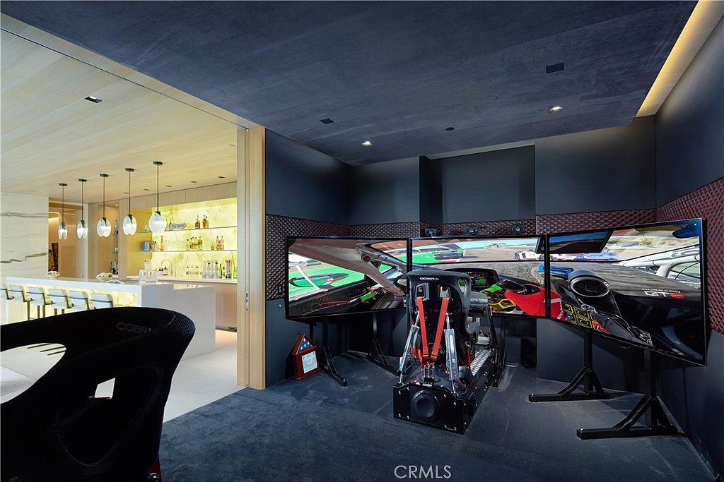 This image showcases a high-end home gaming room featuring a professional-grade racing simulator setup with three large monitors and a bucket seat. The room is designed with dark, sound-dampening wall panels and a sleek, modern aesthetic, while an adjacent bar area with illuminated shelving is visible through an open doorway. The perspective is centered on the simulator, highlighting the immersive entertainment space within the home.