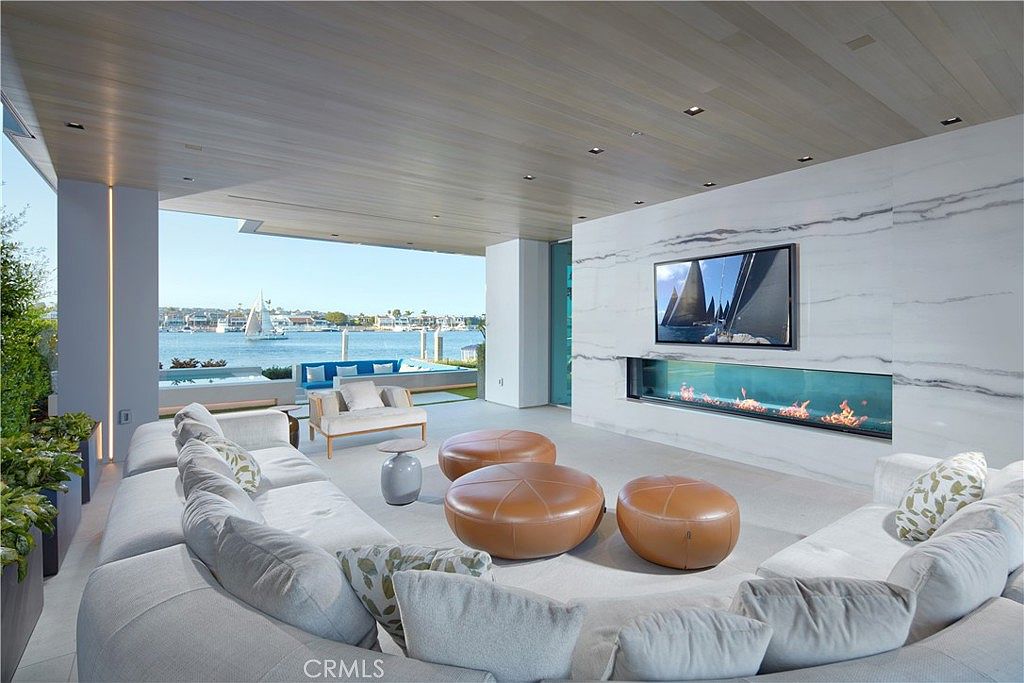 This expansive, open-concept living area seamlessly blends indoor and outdoor living, featuring a large, light-toned sectional sofa and three leather ottomans arranged around a central area rug. A long, linear fireplace is integrated into a prominent stone-clad wall, which also houses a large television. The space opens directly to a waterfront patio, offering an unobstructed view of the harbor and boats through a wide, open-air transition.