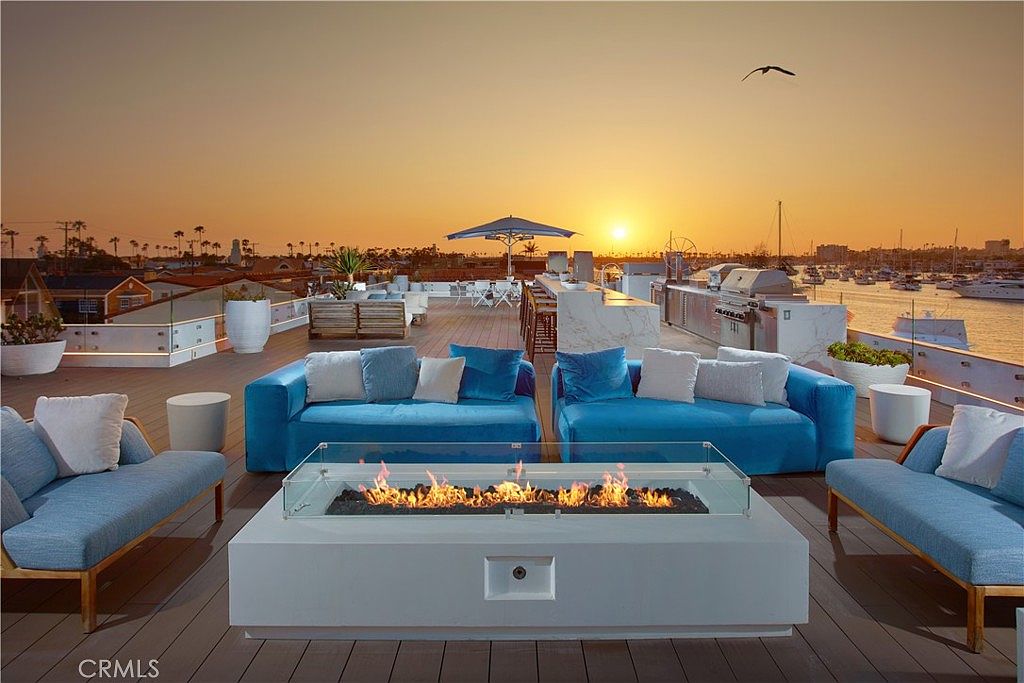 This expansive rooftop deck offers a luxurious outdoor living space overlooking a marina at sunset. The area is furnished with multiple vibrant blue sofas arranged around a central rectangular fire pit with a glass wind guard, complemented by a sleek outdoor kitchen and bar area. The scene captures a serene, high-end atmosphere with panoramic water views and a clear sky, perfect for evening entertaining.