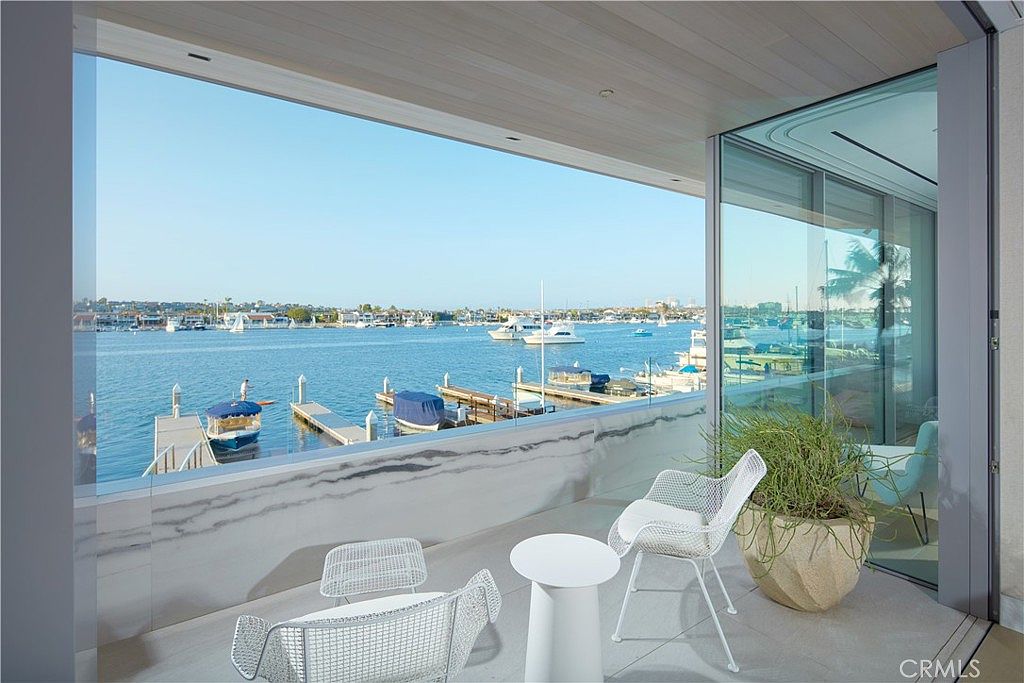 This modern balcony offers a seamless indoor-outdoor transition with floor-to-ceiling glass walls that frame a panoramic view of the harbor. The space is furnished with minimalist white wire chairs and a small round table, set against a sleek stone-clad railing. The perspective captures the serene waterfront setting, emphasizing the open-air design and the tranquil atmosphere of the coastal location.