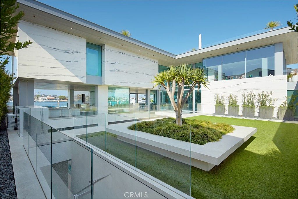 This modern luxury residence features a striking architectural design with extensive glass walls and white stone cladding. The rear exterior showcases a manicured lawn, a central planter with a sculptural tree, and a glass-railed terrace overlooking a waterfront harbor. The perspective captures the seamless integration between the indoor living spaces and the outdoor environment, emphasizing a sophisticated, contemporary aesthetic.