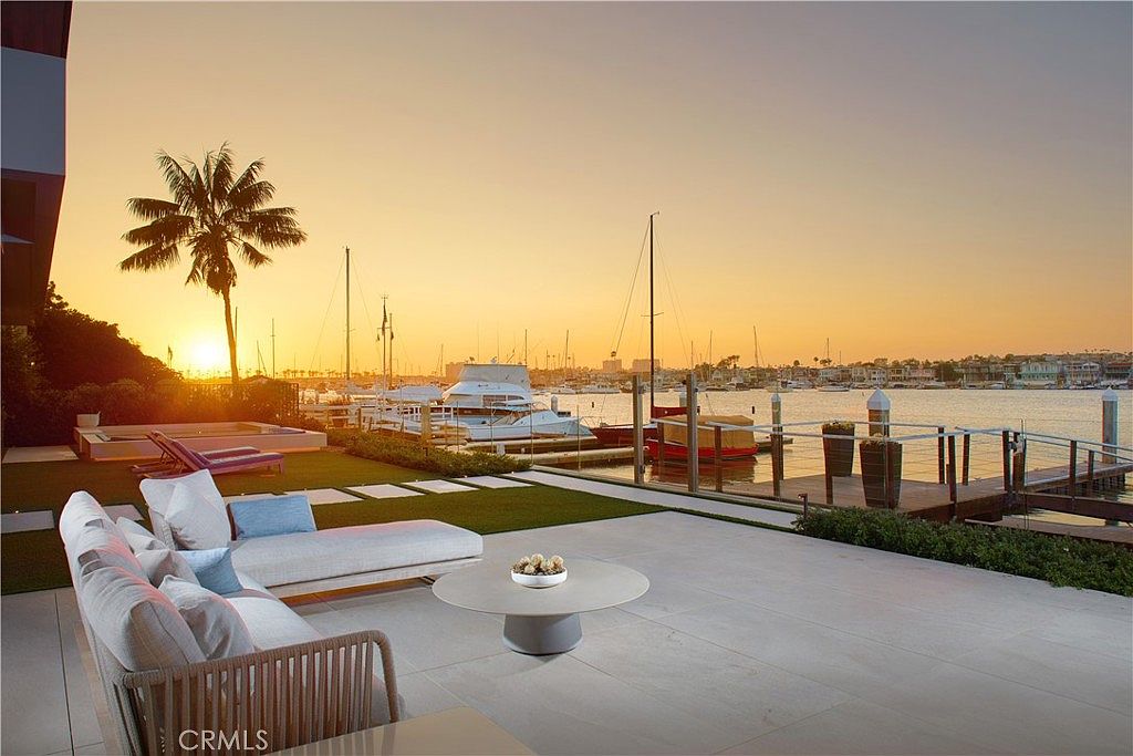 This luxurious waterfront patio offers a serene outdoor living space with a modern sectional sofa and a circular coffee table set against a stunning sunset backdrop. The paved terrace transitions into a manicured lawn area featuring lounge chairs and a small spa, all overlooking a private dock and a calm harbor filled with sailboats. The scene captures an elegant, tranquil atmosphere perfect for evening relaxation by the water.